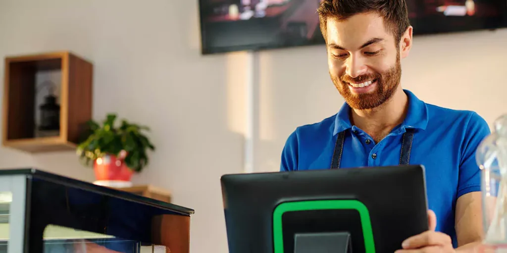 Smiling bearded coffeeshop barista working on POS terminal when entering customer order or accepting payment