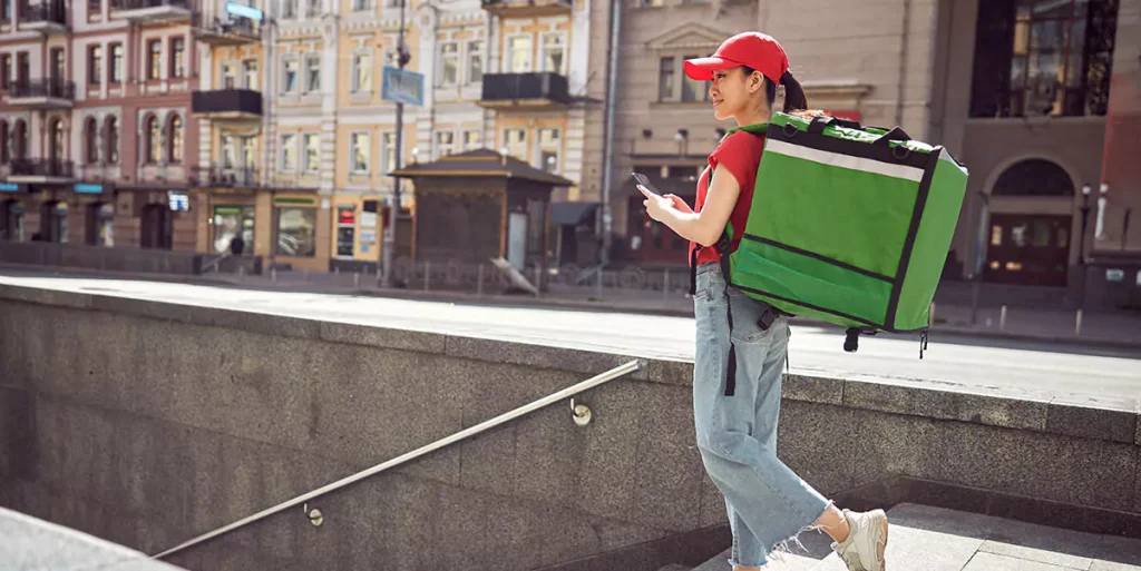 Side view of female courier with green thermo backpack, walking down street for food delivery service during coronavirus pandemic