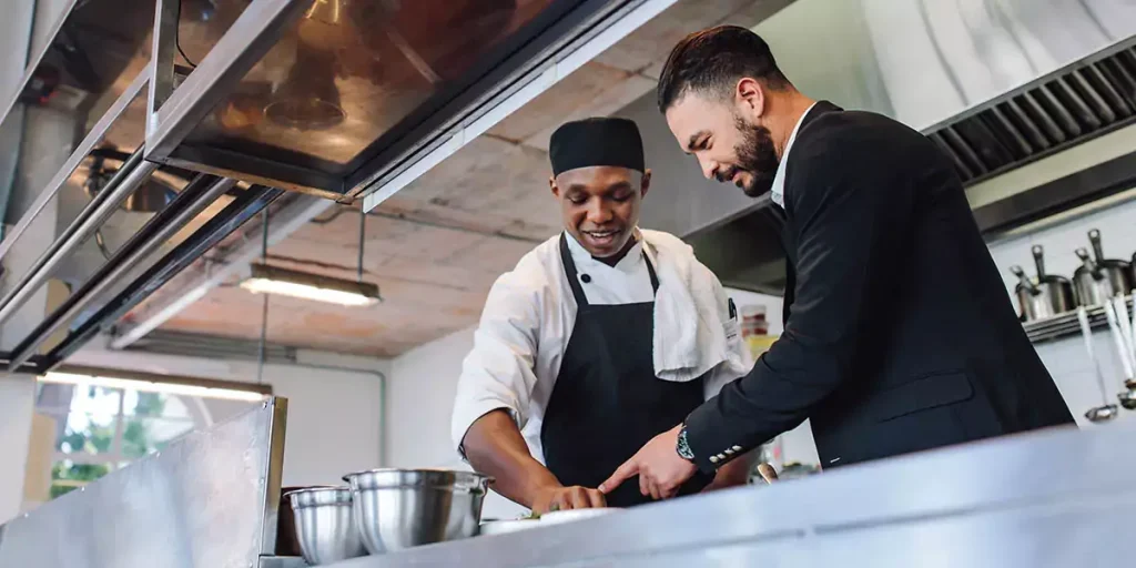 Restaurant manager discussing with chef in kitchen. Cook preparing a dish with restaurant owner standing by.